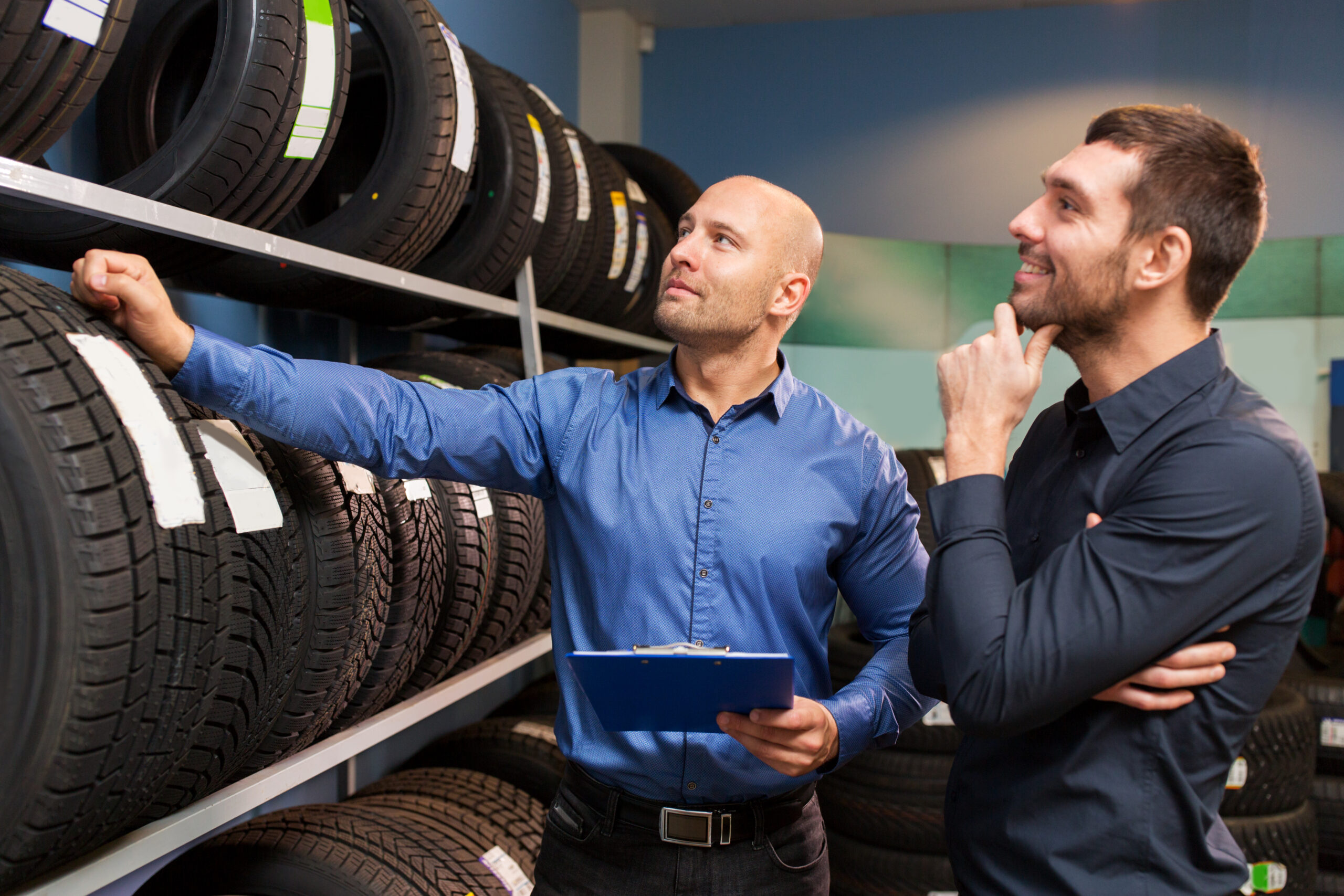 business, maintenance and people concept - male customer and salesman with clipboard showing wheel tires at car repair service or auto store