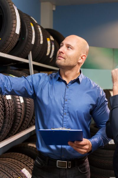 business, maintenance and people concept - male customer and salesman with clipboard showing wheel tires at car repair service or auto store