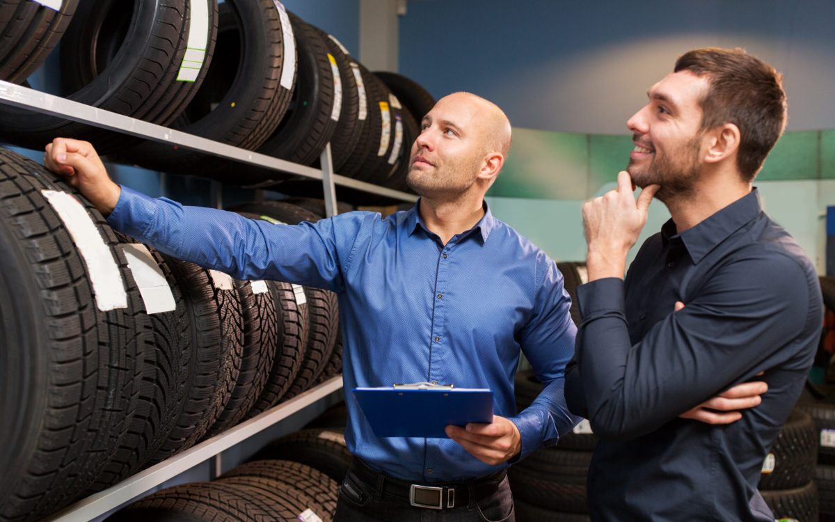business, maintenance and people concept - male customer and salesman with clipboard showing wheel tires at car repair service or auto store